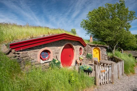Side view of The Burrow with round red door, circular windows and grass-covered roof at Craighead Howfs